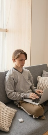 На фото парень сидит на диване у себя в комнате перед ноутбуком занимаясь курсом английского языка